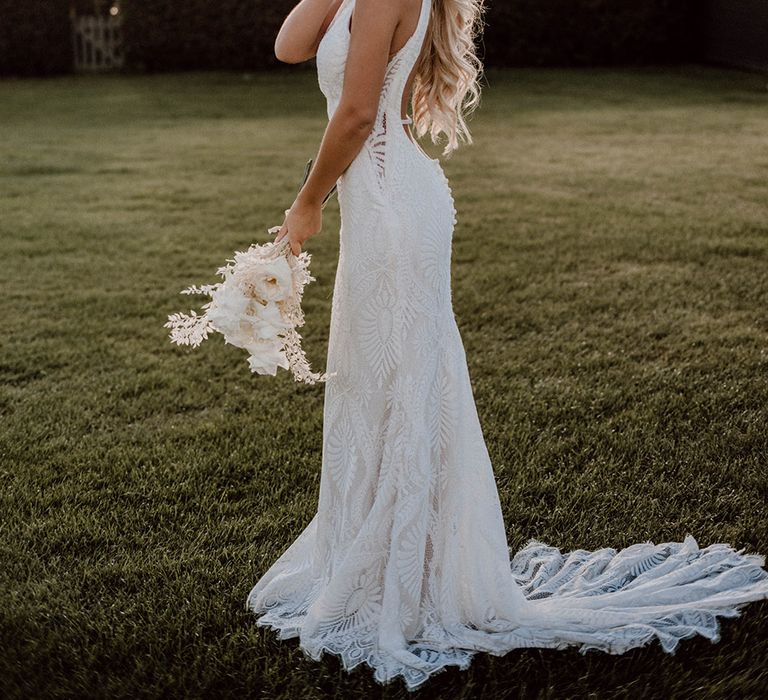 Blonde bride with long hair in a fitted wedding dress with pattern detail and low back design holding a white wedding bouquet 