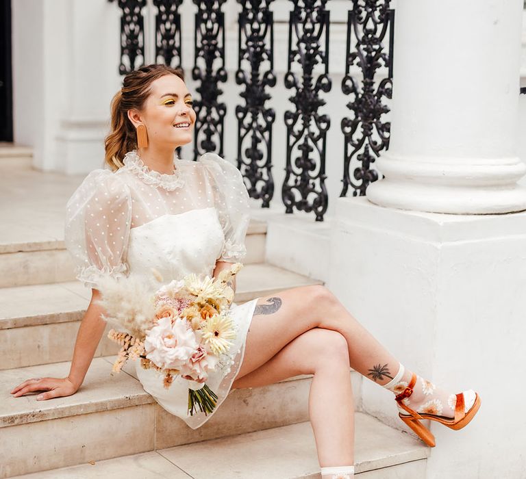 Stylish bride in a polka dot tulle wedding dress, platform sandals and floral socks holding a yellow wedding bouquet 