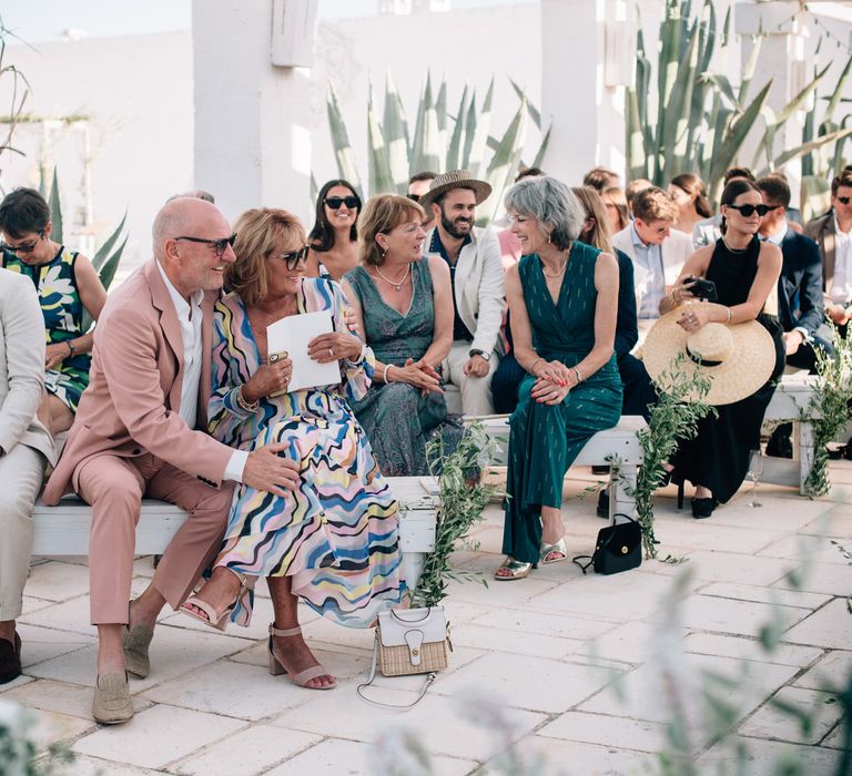 Stylish wedding guests sitting on white benches for rustic outdoor Italian wedding ceremony