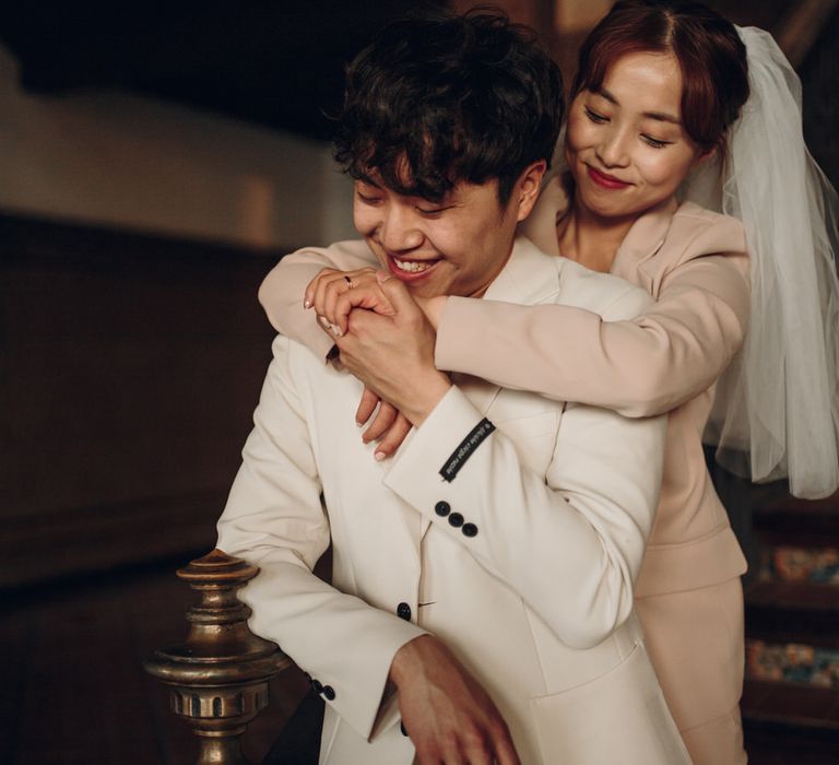 Asian woman wearing veil stands behind partner wearing cream suit