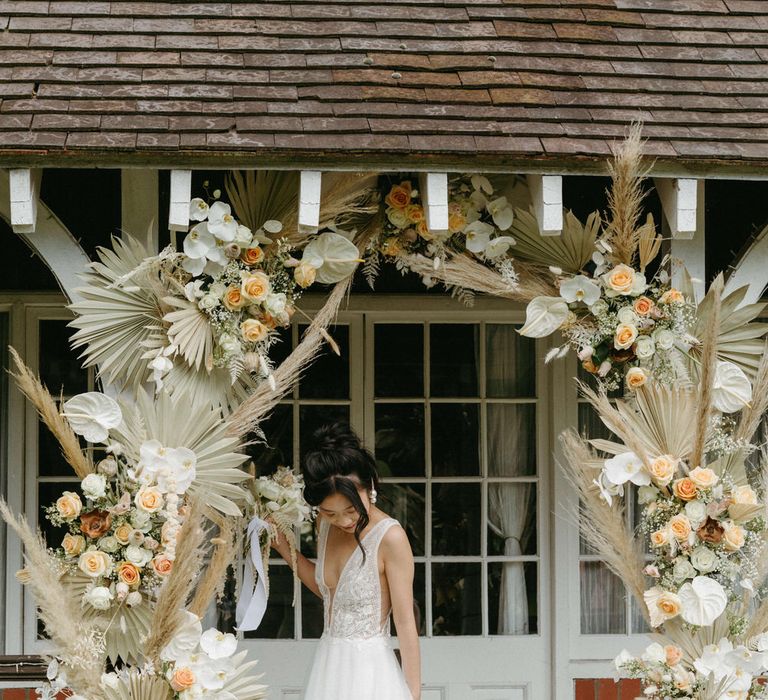 Bride at Berwick lodge wearing a plunge neck wedding gown with tulle skirt, under an arch of dried palms, pampas grass, white anthuriums, roses and orchids