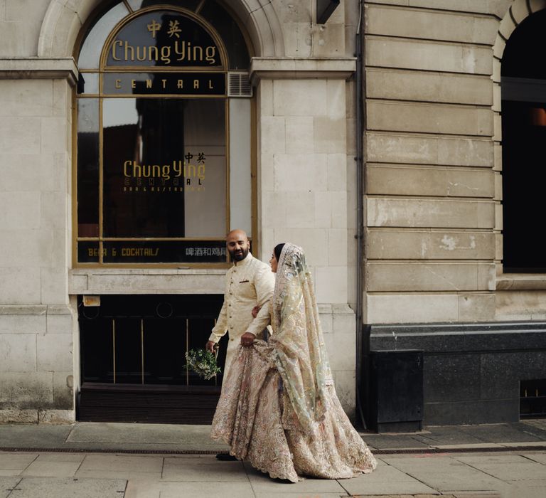 Bengali groom in a gold coat helping his bride with her pastel Suffuse by Sana yasir wedding dress as they walk through the streets of Birmingham