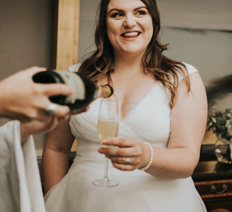 Bride has champagne poured into flute as she smiles and wears pearl bracelet 