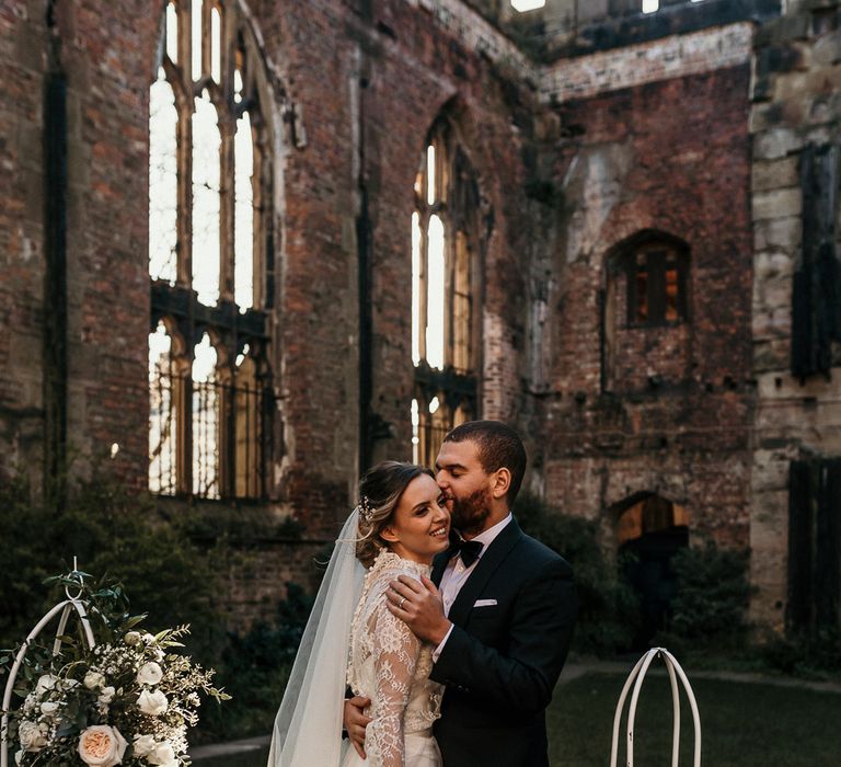 Groom in a black tuxedo kissing his brides head in a lace and tulle fitted wedding dress at St Luke's Bombed Out Church in Liverpool 