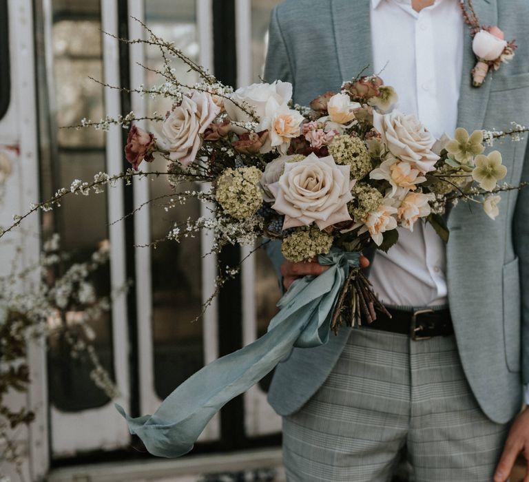 Grooms wears pale blue suit with tartan trousers whilst carrying floral bouquet 