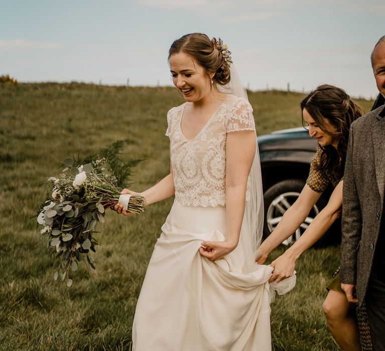 Bride in lace top wedding dress with capped sleeves and satin skirt holds white and green mixed bouquet whilst walking through field with wedding guest holding train before Dunluce Castle wedding