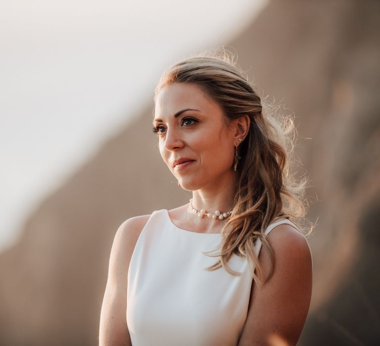 Bride looks out toward the ocean as she wears her blonde hair in loose curls with vintage pearl necklace