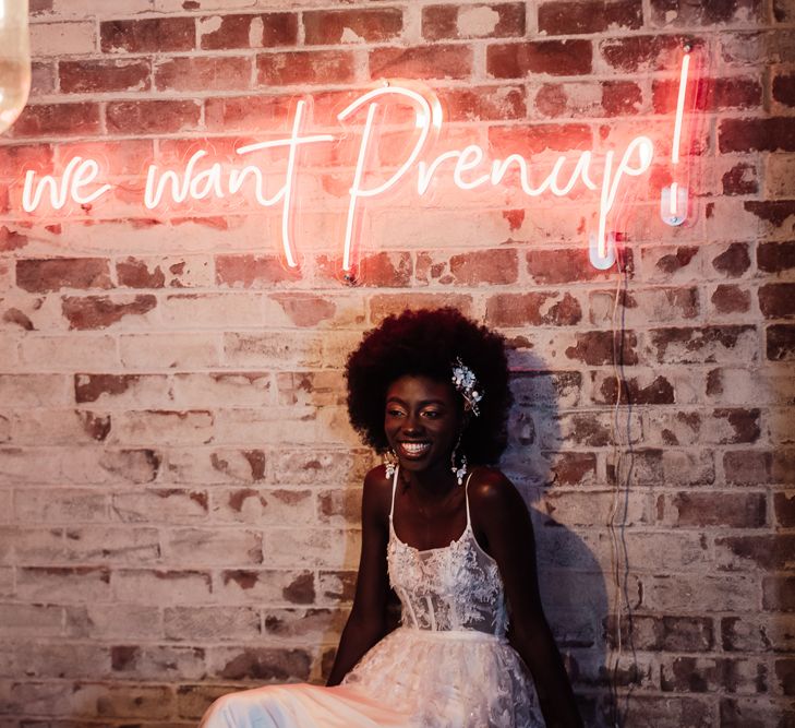 A Black bride with natural afro sits laughing in a white wedding dress in front of a neon sign that reads holla we want prenup.