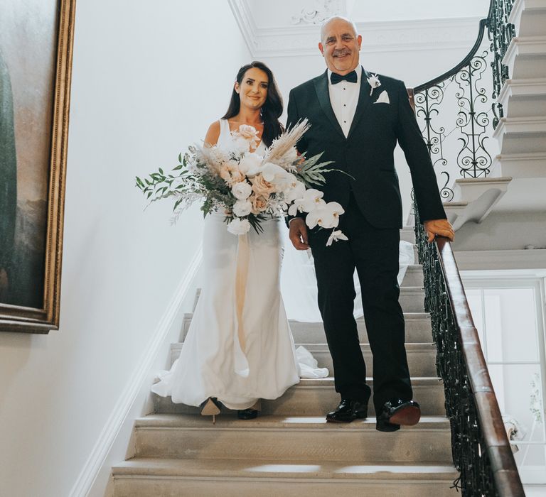 Bride with long dark curled wedding hair wearing a white Pronovias wedding dress with train walks down stairs arm in arm with man in tuxedo as she holds wedding bouquet 