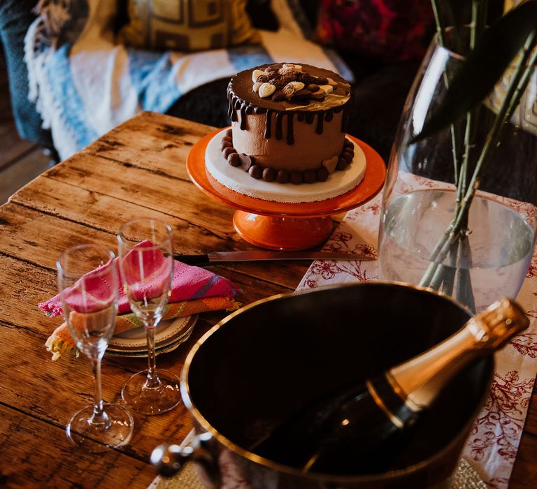 Chocolate wedding cake featuring chocolate fondant dripping around sides sat on wooden table with champagne to the side