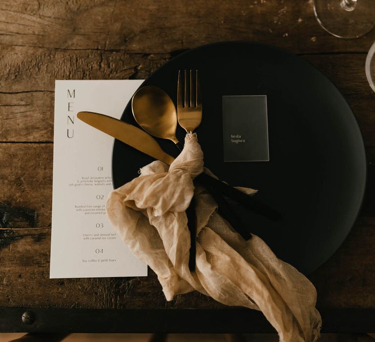 Black tableware with white wedding menu, gold cutlery and neutral linen napkin