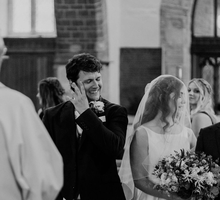 Smiling groom wipes tear from his eye standing next to bride in veil holding white and green bridal bouquet at the altar of church wedding ceremony