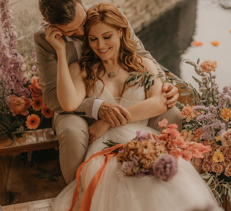 Bride with long wavy red hair sitting on a boat with her groom surrounded by pastel orange, pink and lilac wedding flowers at Euridge Manor boat house 