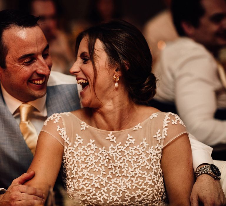 Groom in blue waistcoat and gold tie smiles at laughing bride in lace top Rime Arodaky wedding dress with pearl drop earrings as they sit at wedding breakfast in Dorset