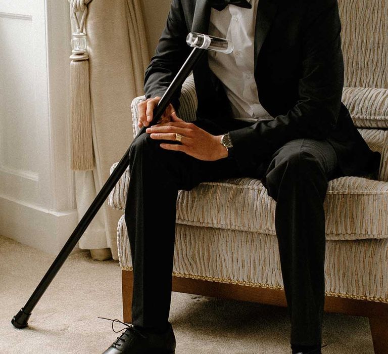 Interable groom in a black tuxedo sitting on a chair at Bourton Hall with his walking stick 