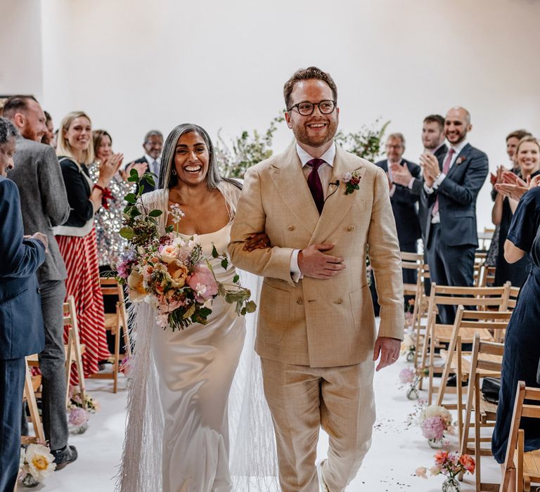 Bride in satin Halfpenny London wedding dress and tasselled bridal cape holding multicoloured bridal bouquet smiles and she walks back down the aisle with groom in double breasted linen suit with gold Nike Air Force 1 trainers after wedding ceremony at Loft Studios London