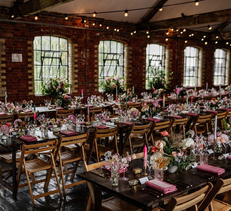 Three long wooden tables with folding wooden chairs, multicoloured candles, mixed florals and pink napkins in industrial room with festoon lighting for wedding breakfast at Loft Studios London