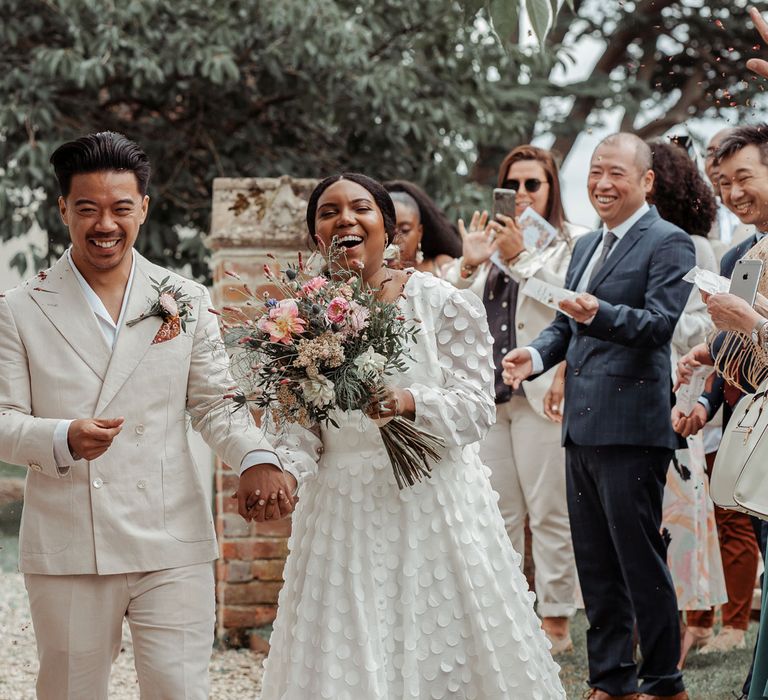 Groom in white shawl collared shirt and double breasted linen suit holds hands with bride in long mesh sleeve spotted Vagabond wedding dress holding mixed bridal bouquet as wedding guests throw confetti at Wasing Park wedding