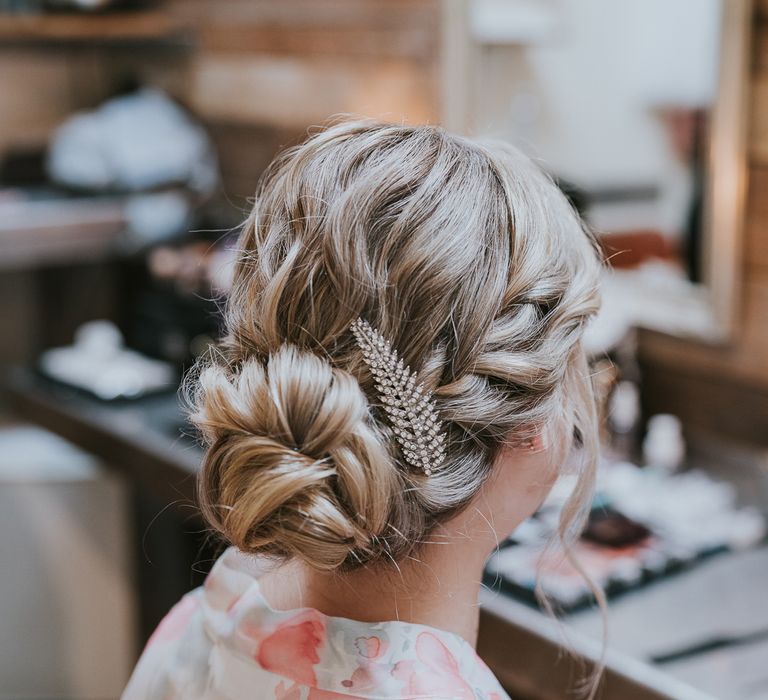 Bride with plaited bun and leaf hair clip sits at mirrors before Tythe Barn wedding with barn wedding flowers