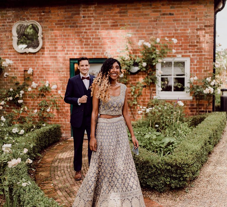 Bride wears traditional lehenga as she walks down pathway as her groom follows from behind | Joshua Gooding Photography