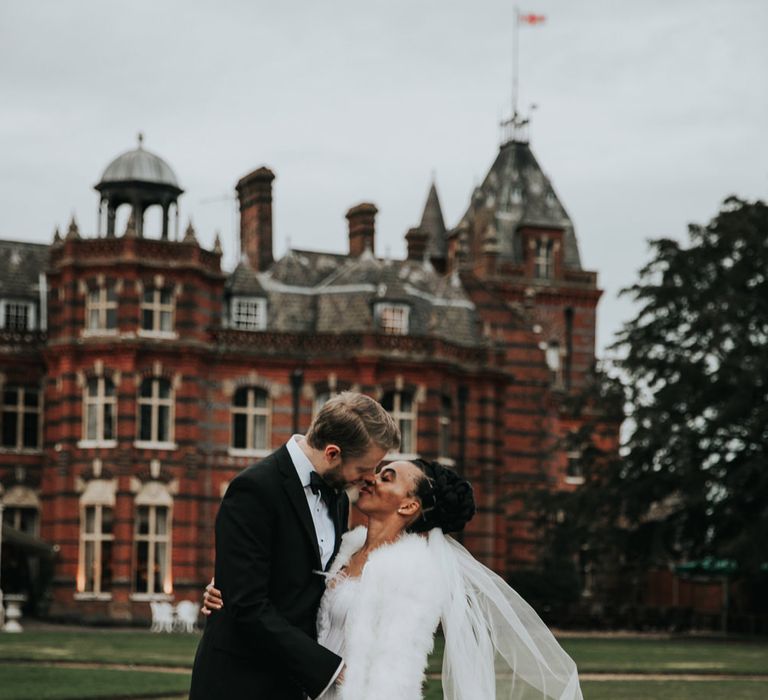 Luxury wedding at The Elvetham with groom in a tuxedo kissing his Black bride in white fluffy bridal cover up 