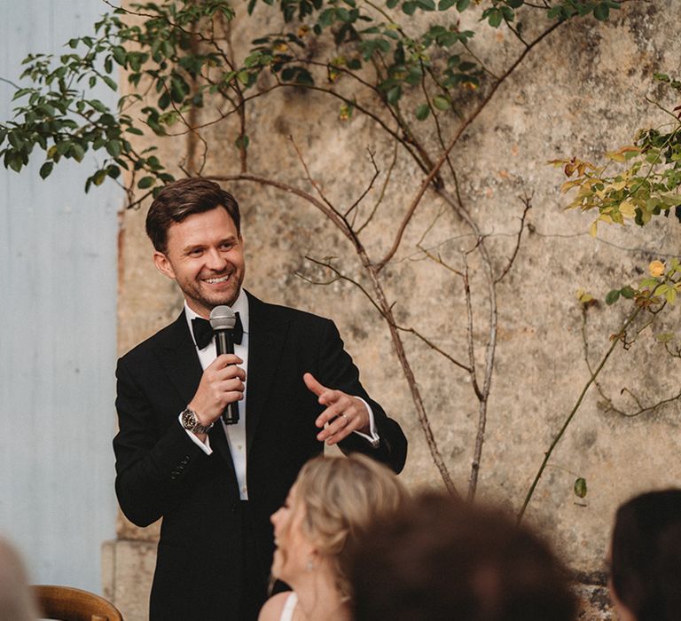 Groom in a tuxedo and bow tie giving a speech at the outdoor wedding reception 