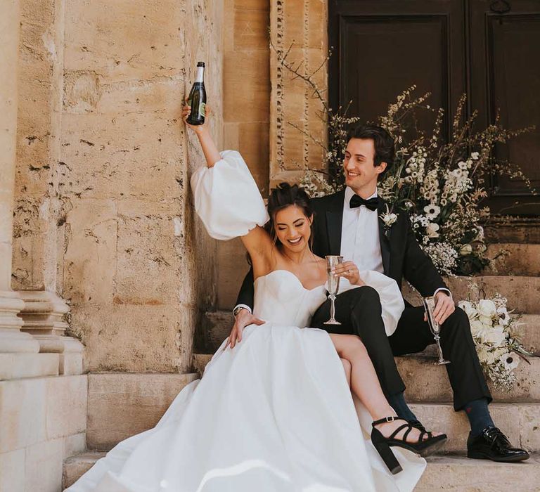 Bride and groom sipping champagne on the steps at Bodleian Library in a tuxedo and Nortier Shallow wedding dress