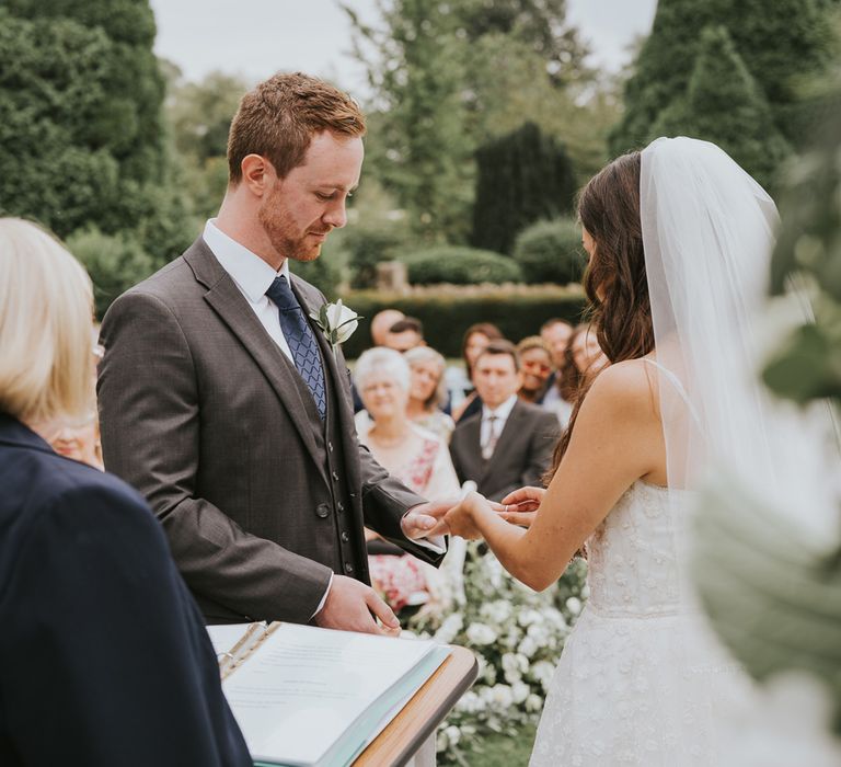 Bride in white Pronovias wedding dress and veil puts ring on hand of groom in grey three piece suit, blue tie and white rose buttonhole during outdoor wedding ceremony