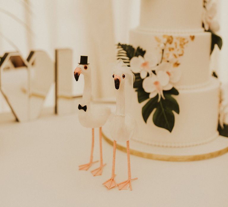 4-tier tropical wedding cake with white frosting, gold leaf decor and flamingo statues