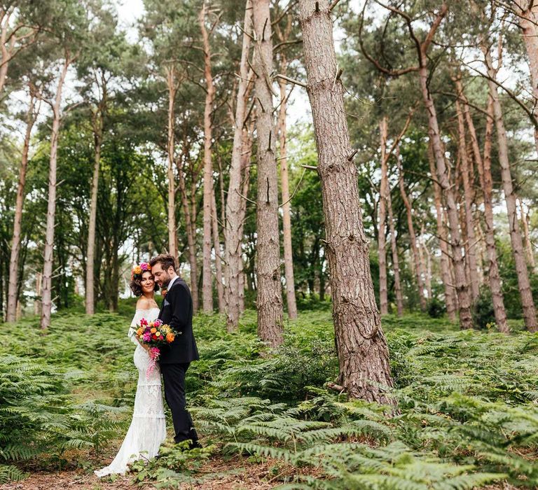 Bride in off the shoulder Grace Loves Lace wedding dress and flower crown holding colourful bridal bouquet stands with groom in black suit in woodland after outdoor wedding ceremony at The Bridal Barn in Claverley