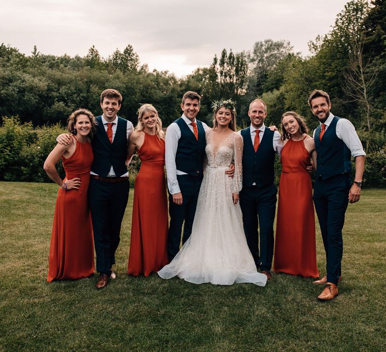 Bride in long mesh sleeve Julie Vino wedding dress stands with bridesmaids in red bridesmaid dresses and groomsmen in dark waistcoats and red ties at tipi wedding reception