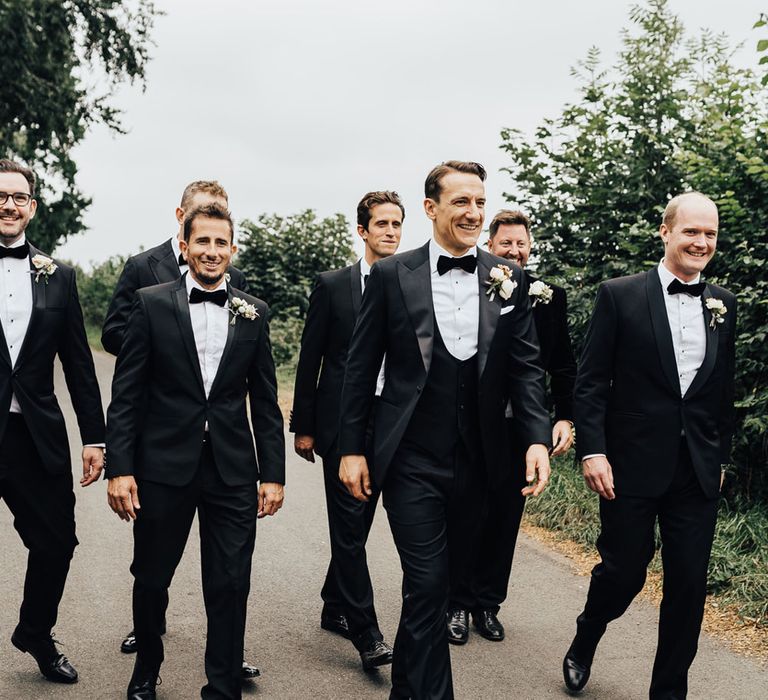 Groom and groomsmen walk down road all dress in black tuxedos with black bow ties and floral buttonholes before church wedding ceremony in Hertfordshire