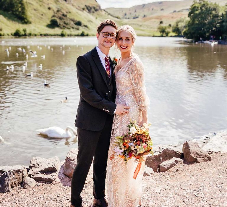 Bride & groom stand with their arms around one another in front of lake on their wedding day