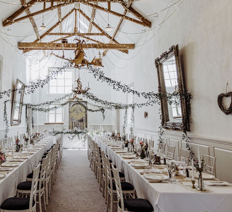 Interior of stone barn wedding reception with long tables, bamboo style chairs, floral garlands and fairy lights for rustic chic 