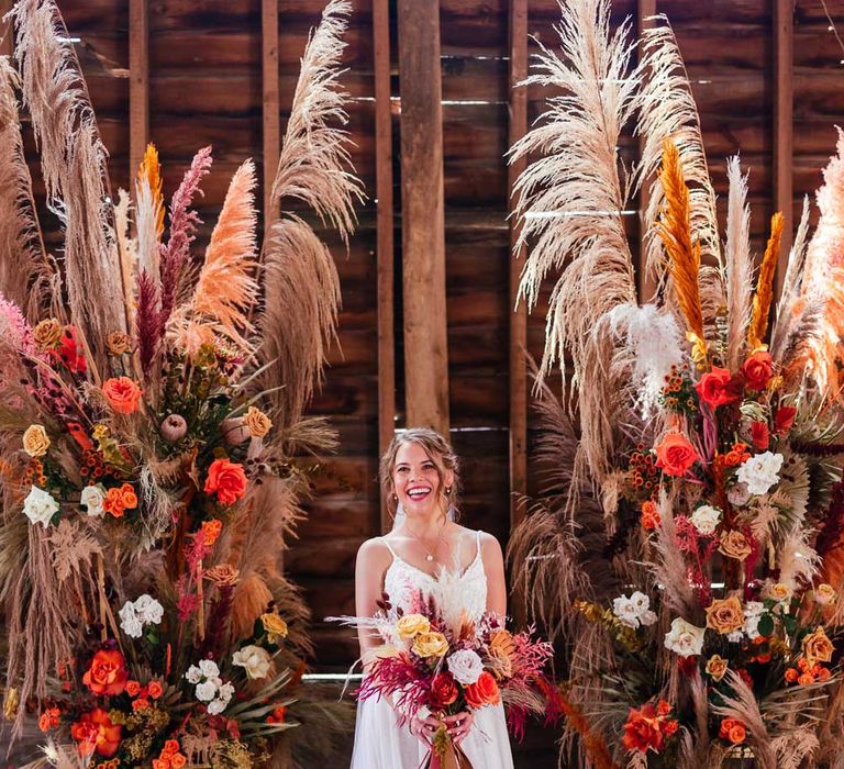 Smiling bride in lace thin strap wedding dress with train holds white and red rustic bridal bouquet and stands by rustic floral columns with pampas grass and roses for festival style wedding