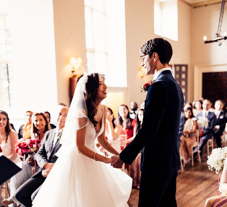 Bride in layered tulle wedding dress and veil stands holding hands with groom in dark suit and floral buttonhole during Elmore Court wedding ceremony