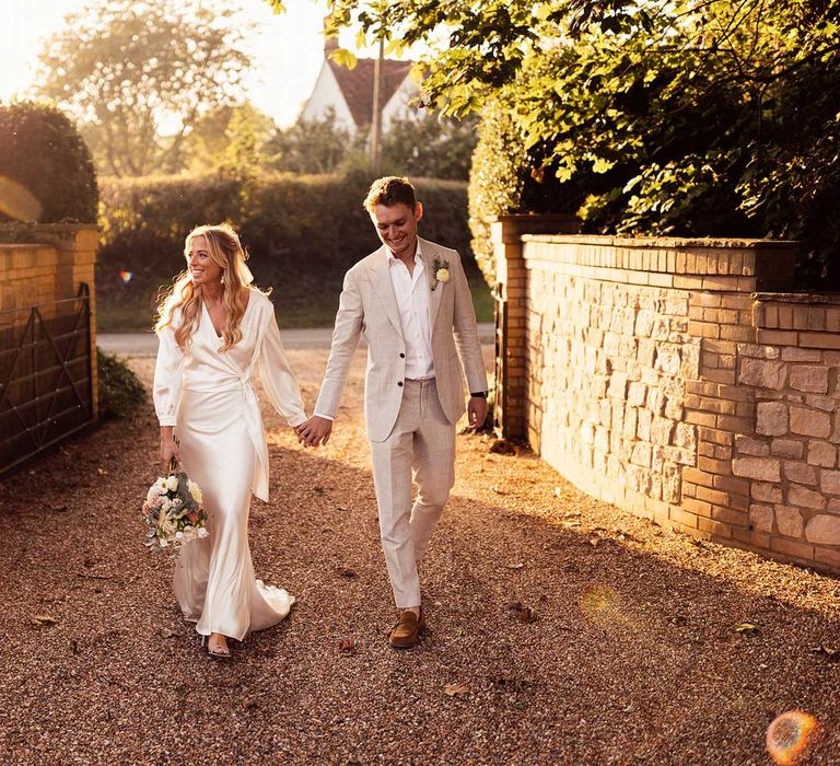 Smiling bride in satin wedding dress and wrap top walks down drive holding hands with groom in grey suit and open white shirt