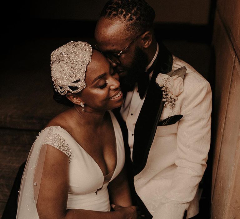 Groom in a white tuxedo jacket kissing his bride in a beaded cap vintage style wedding headdress at their vintage style city wedding 