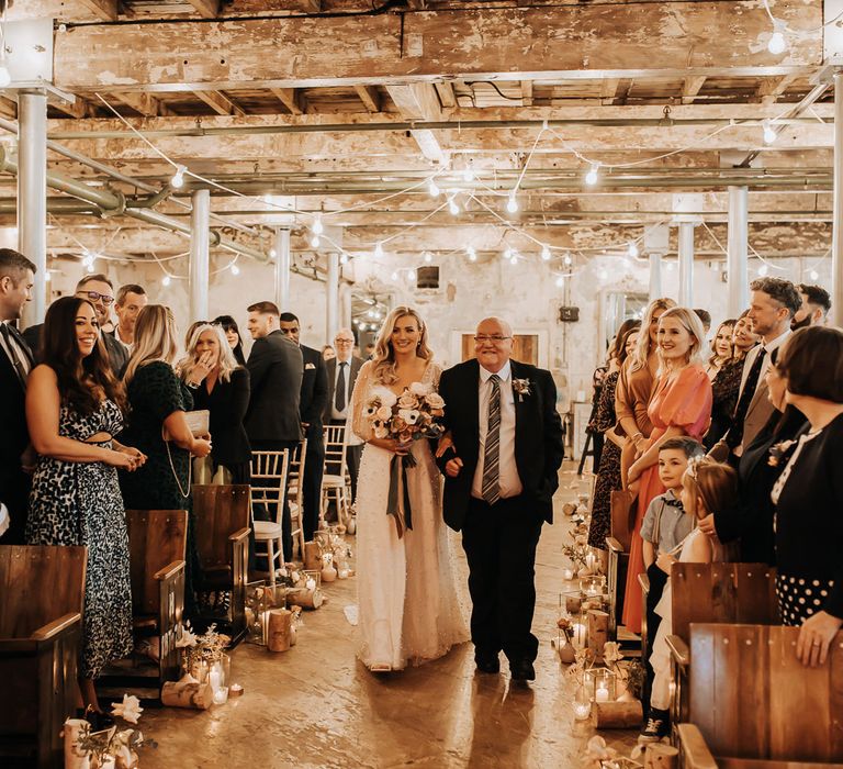 Bride in white pearl wedding dress with sleeves and pastel mixed bouquet with roses walks down the aisle with man in dark suit and striped tie at industrial style wedding