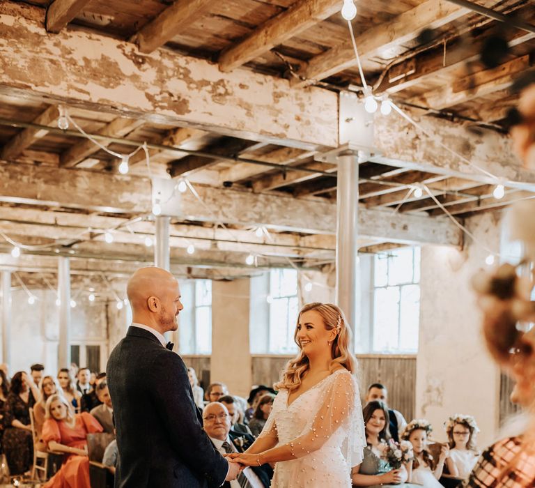 Bride in white v neck wedding dress with pearls stands with groom in navy suit during industrial style wedding ceremony