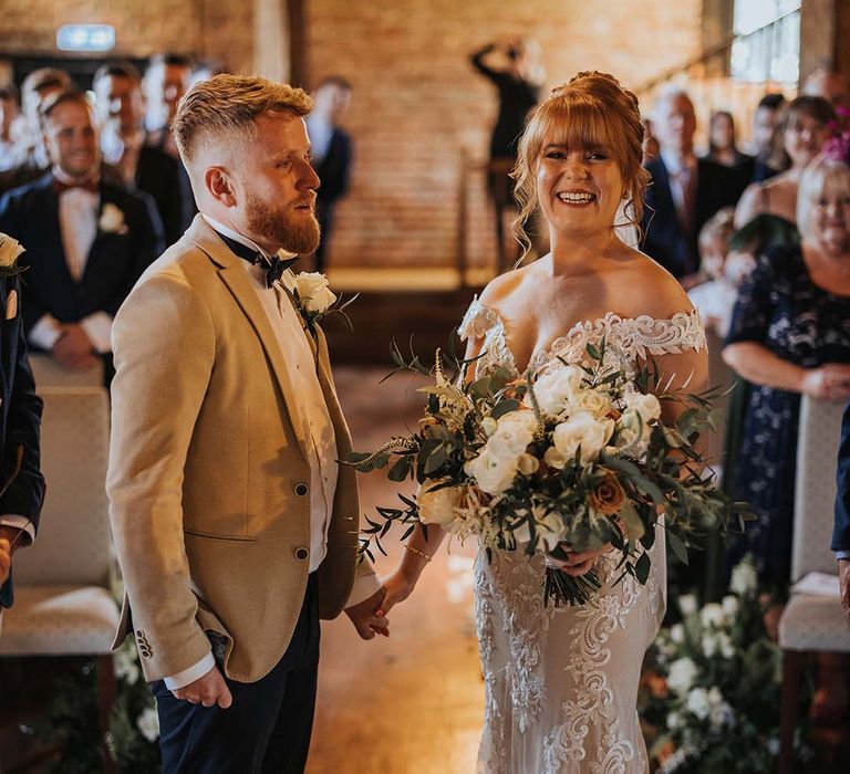 Groom in beige suit jacket with bow tie stands with bride at the end of the altar with bride in Maggie Sottero lace wedding dress