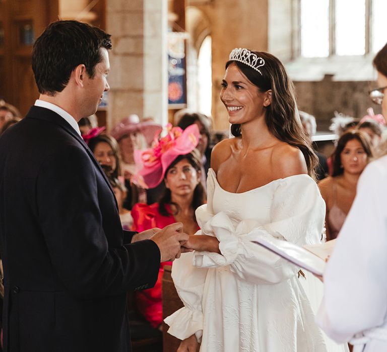 Groom places on wedding and to bride's finger as bride wears off the shoulder puff sleeve wedding dress with tiara crown