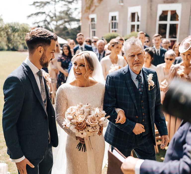 Father of the bride in blue checkered suit gives away bride in beaded dress to groom in dark blue suit 