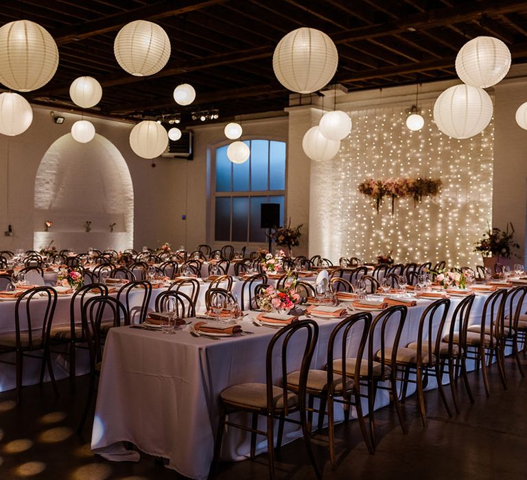 Wedding venue with white hanging paper lanterns and long tables for reception with fairy lights and a pink flower cloud