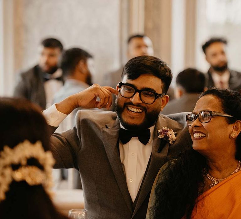 Groom in dark grey suit and black bow tie embracing wedding guest in vibrant orange saree 