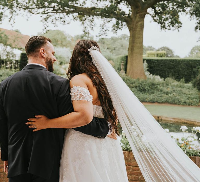 Groom in dark grey jacket hugs bride in lace off the shoulder dress and veil looking out over the water 