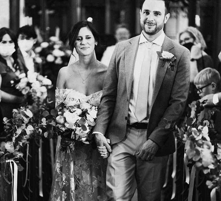 Bride and groom hold hands as they walk back down the aisle as a married couple with bride wearing lace wedding dress and groom in grey suit