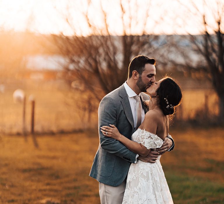 Bride and groom share a kiss in fields as the sun begins to set