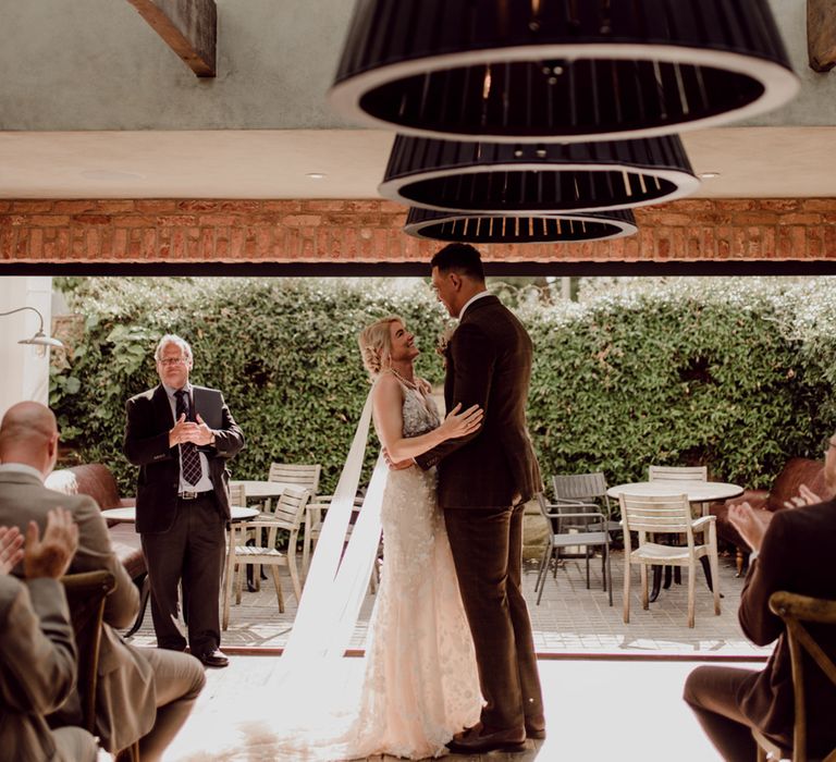 Groom in brown suit smiles down at bride in lace wedding dress with pearl necklace