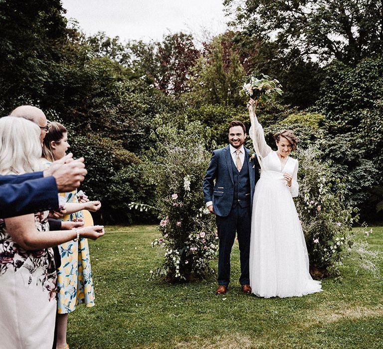 Bride in long sleeve wedding dress raises her bouquet in celebration with groom in blue suit at the altar